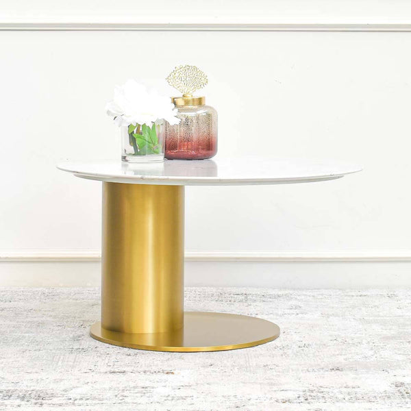 A gold stainless steel coffee table with a sleek, minimalist design, showcasing a sculptured curved base and a white sintered stone tabletop, accessorized with a decorative jar and flowers.