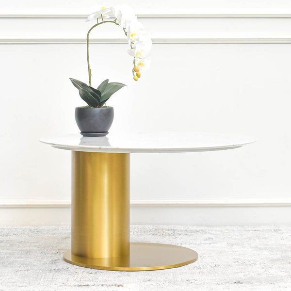 A gold steel coffee table featuring a minimalist design with a sculptured curved base and a white sintered stone tabletop, embellished with an orchid plant.
