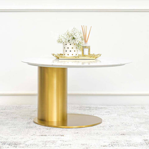 A gold stainless steel coffee table with a sculptured curved base and a white sintered stone tabletop, adorned with a vase of white flowers and a decorative tray.