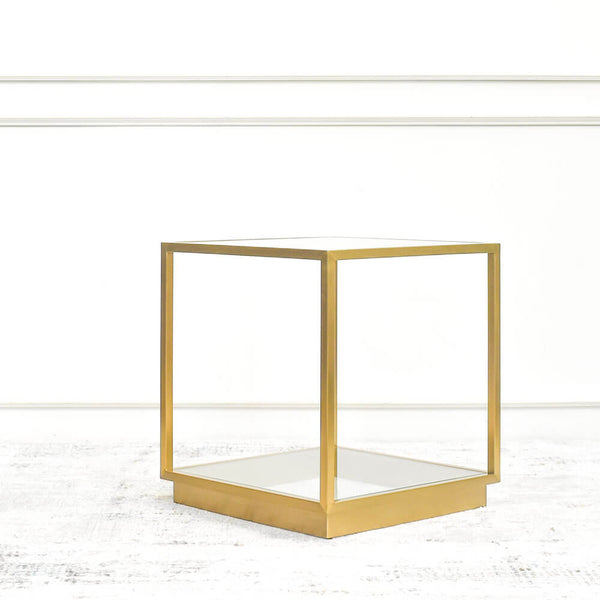 A gold side table with a minimalist design, featuring a square frame and a glass tabletop, displayed at a living room set-up in a luxury furniture showroom.