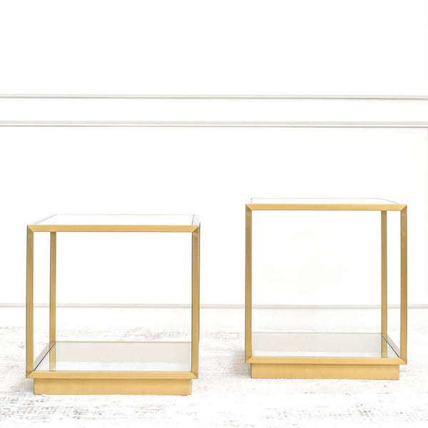 Two gold steel side tables with square frames and glass tabletops, placed on a white floor against Finn Avenue furniture showroom wall.