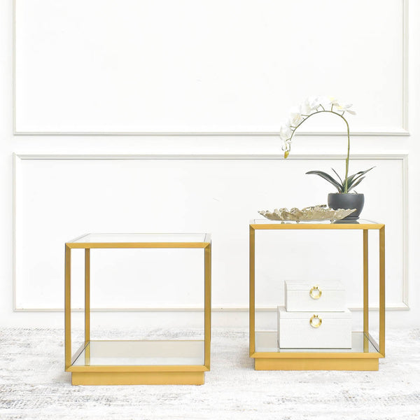 Two gold square side tables with minimalist designs, showcasing glass tabletops and metal frames, placed on a luxury area rug against white living room wall.