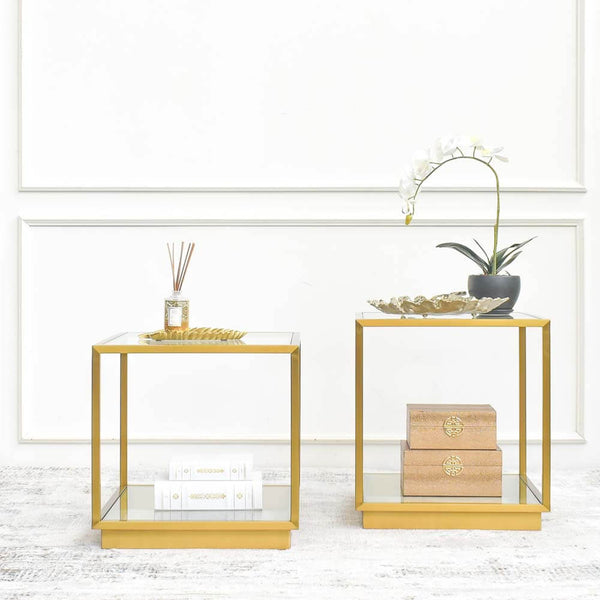 A set of gold stainless steel side tables with clear glass tabletops, one adorned with a golden leaf-shaped tray containing Finn Avenue reed diffuser. Both are placed on a luxury rug against a white feature wall.