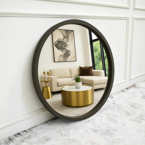 A large, round, charcoal-gray mirror leans against a white paneled wall, reflecting a modern luxury living room with a beige sectional sofa, gold accent table, and a gold-and-white marble coffee table. A black-framed window and abstract wall art add to the elegant, minimalist aesthetic.