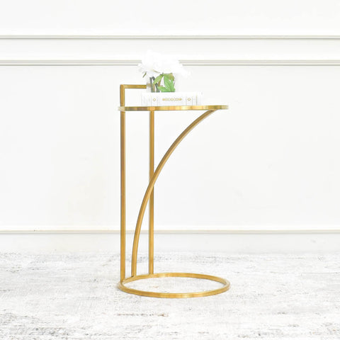 A single gold plated stainless steel side table with a circular tier, featuring a small white flower vase and a stack of books on top. The table is positioned against a white wall with decorative paneling.