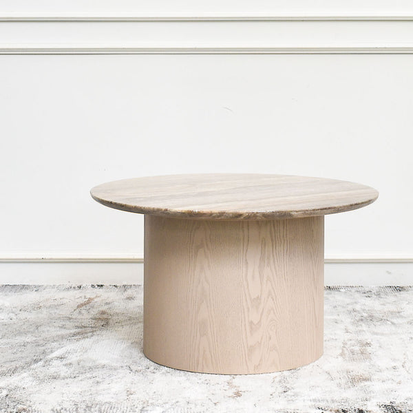 Seroja round travertine marble coffee table with a light wooden base. The neutral tones complement the simple background.