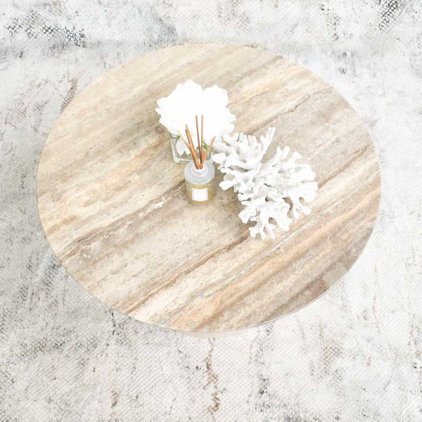 A round coffee table with a travertine marble top and neutral decor, featuring a white coral sculpture, Autograph reed diffuser, and white flowers.