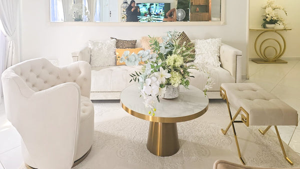 The elegant Duchess Ivory Chesterfield Sofa from Finn Avenue, adorned with decorative cushions including the Plumes Leaves Cushion, anchors a luxurious living room in Singapore. This affordable luxury sofa is beautifully paired with a Raphael Armchair, a Marq Stool with a gold base, and a stylish coffee table, showcasing a sophisticated design by Finn Avenue.