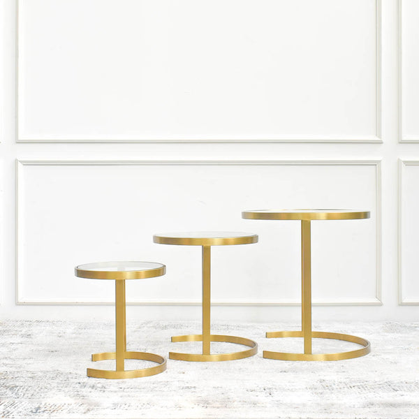 Three gold side tables with circular tiers, arranged in ascending order of height, against a white wall with decorative paneling.