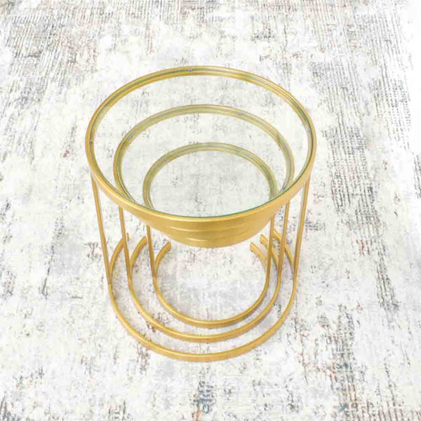 A set of three circular tiered gold side tables with clear glass table tops, arranged in ascending order of height, placed on area rug.