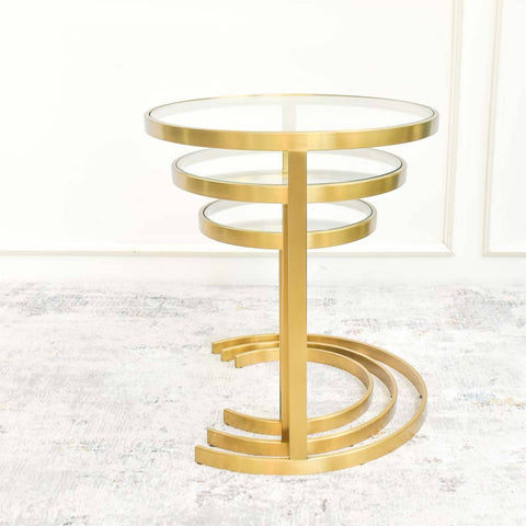 A gold plated stainless steel side table nested in three circular tiers, placed against a white wall on a Finn Avenue luxury rug.